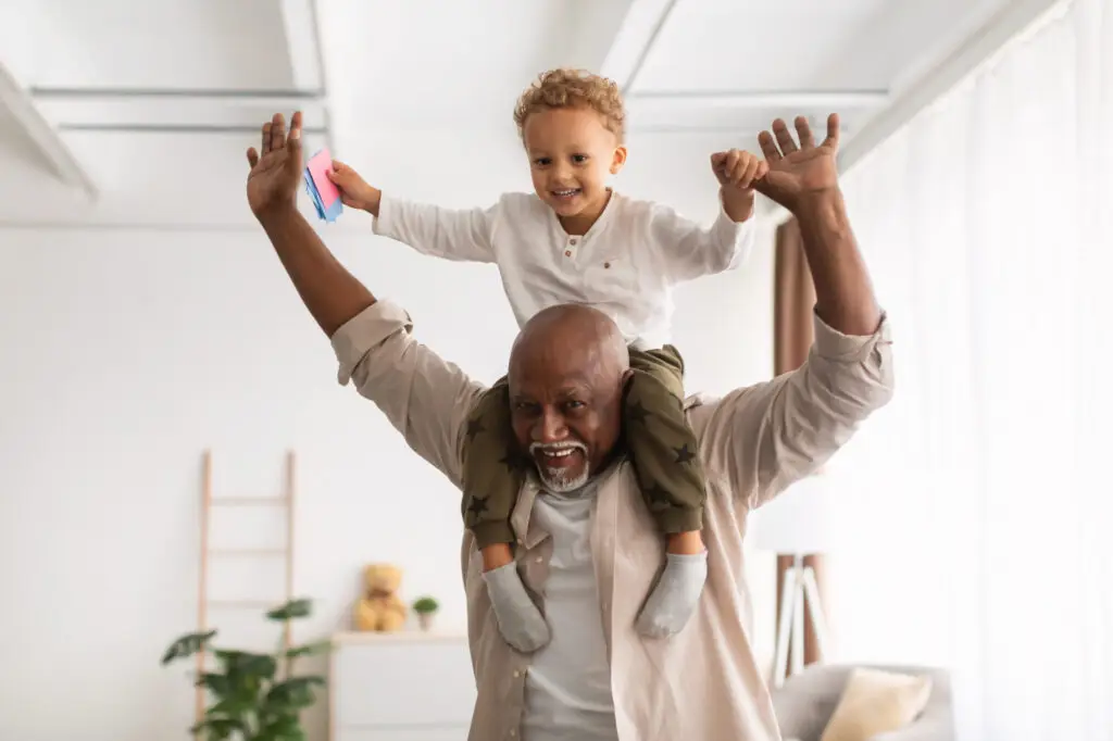 Grandpa Carrying Little Grandson On Shoulders Playing And Having Fun Together At Home.